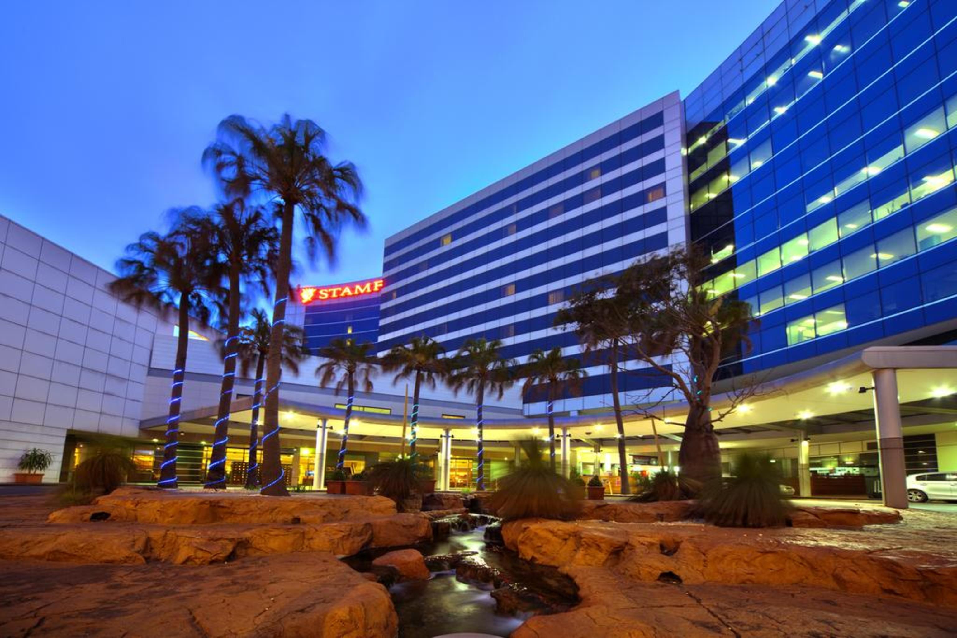 Stamford Plaza Sydney Airport Hotel & Conference Centre Exterior photo The photo shows a modern building, likely a hotel or commercial structure, illuminated during twilight. The facade features a combination of glass and concrete, with blue-tinted windows. In the foreground, there are several palm trees and rocks, sugg