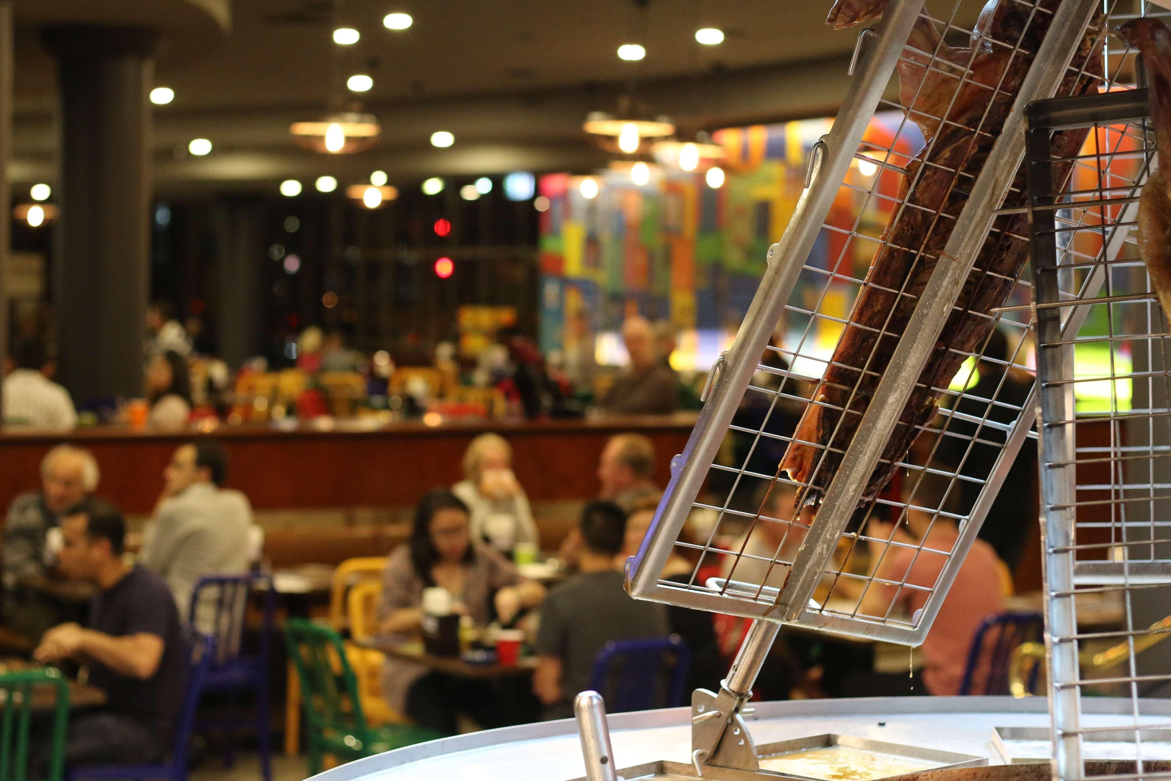 Stamford Plaza Sydney Airport Hotel & Conference Centre Exterior photo The photo shows a bustling indoor dining area with a number of people sitting at tables. In the foreground, there is a grill or cooking apparatus with a grid-like structure, possibly used for preparing food. The background features colorful decoratio